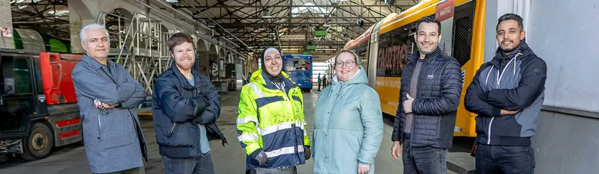 Sechs Menschen, teilweise in Arbeitskleidung in einer großen Halle nebeneinander stehend