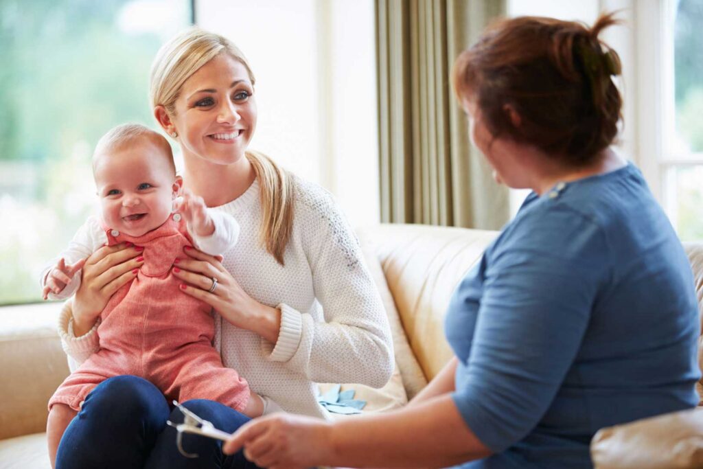 Eine junge Frau sitzt mir Ihrem Kleinkind auf einem Sofa und ist im Gespräch mit einer Mitarbeitenden