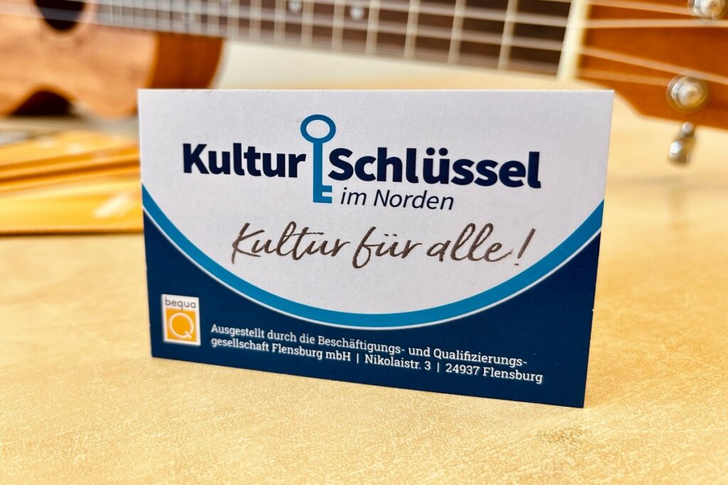 Visitenkarte des Kulturschlüssel Flensburg steht auf einem Tisch; im Hintergrund eine Gitarre